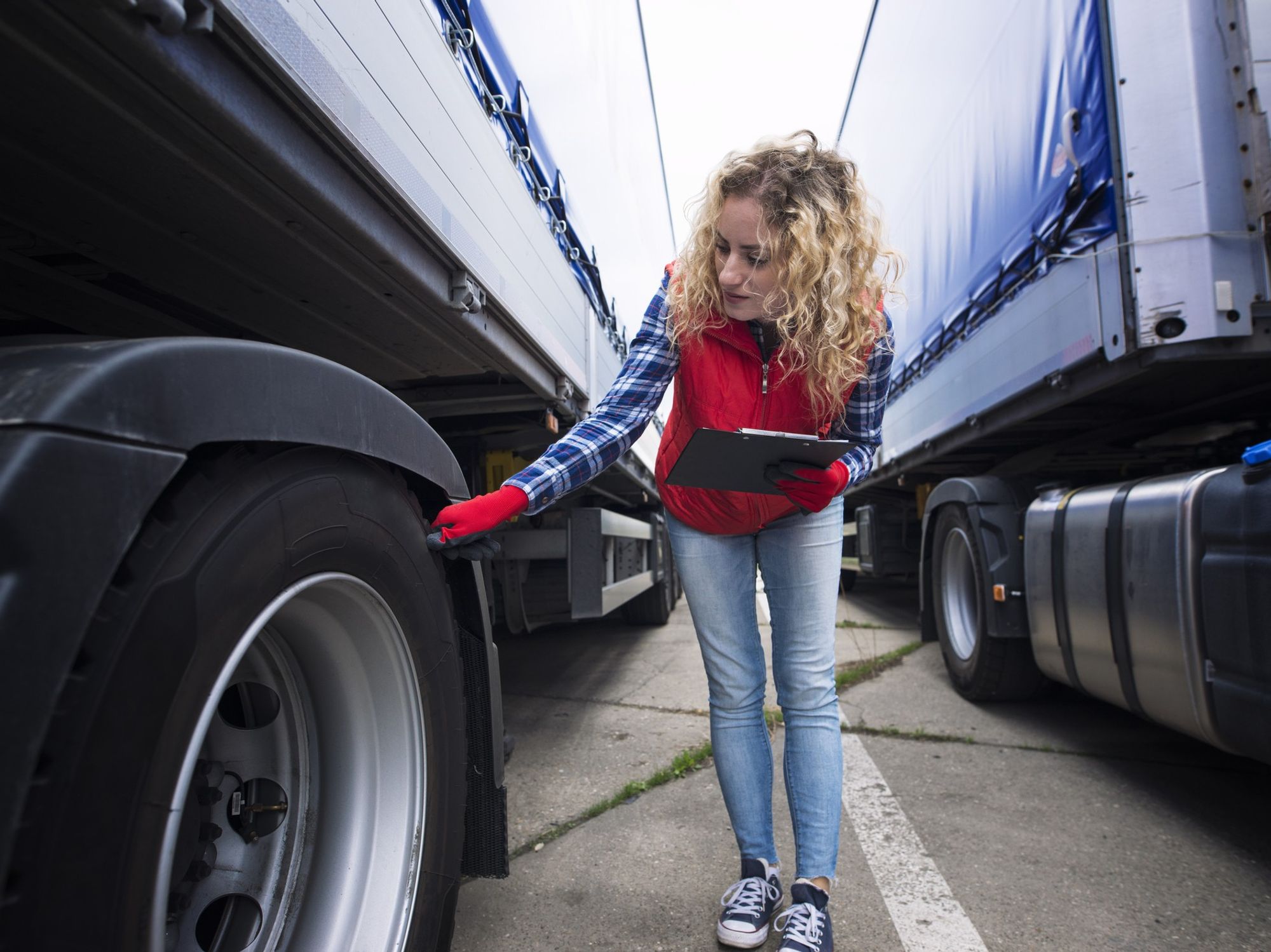 A woman holds a clipboard and checks the tire of a semi-truck.