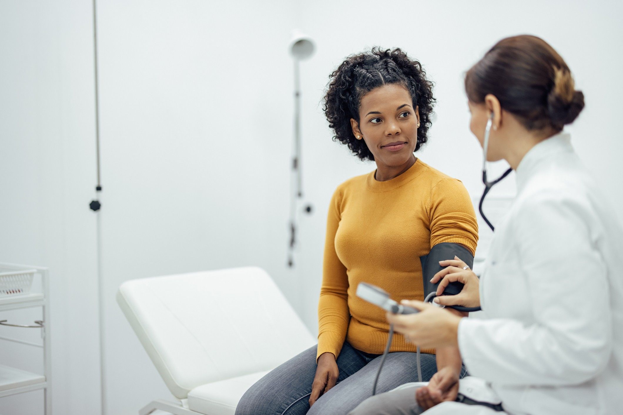 A woman in a yellow shirt sits on an exam table while a doctor in a white coat takes her blood pressure.
