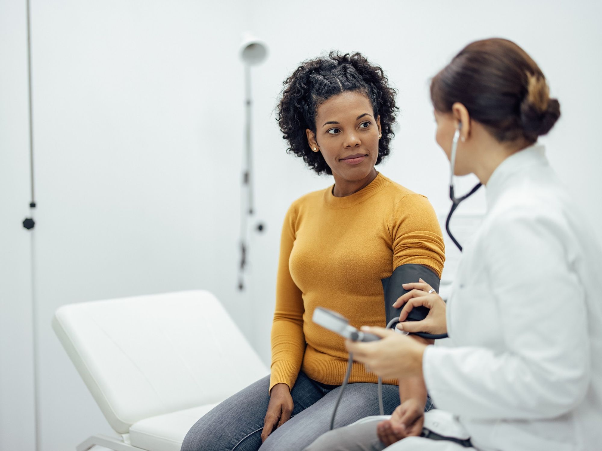 A woman in a yellow shirt sits on an exam table while a doctor in a white coat takes her blood pressure.