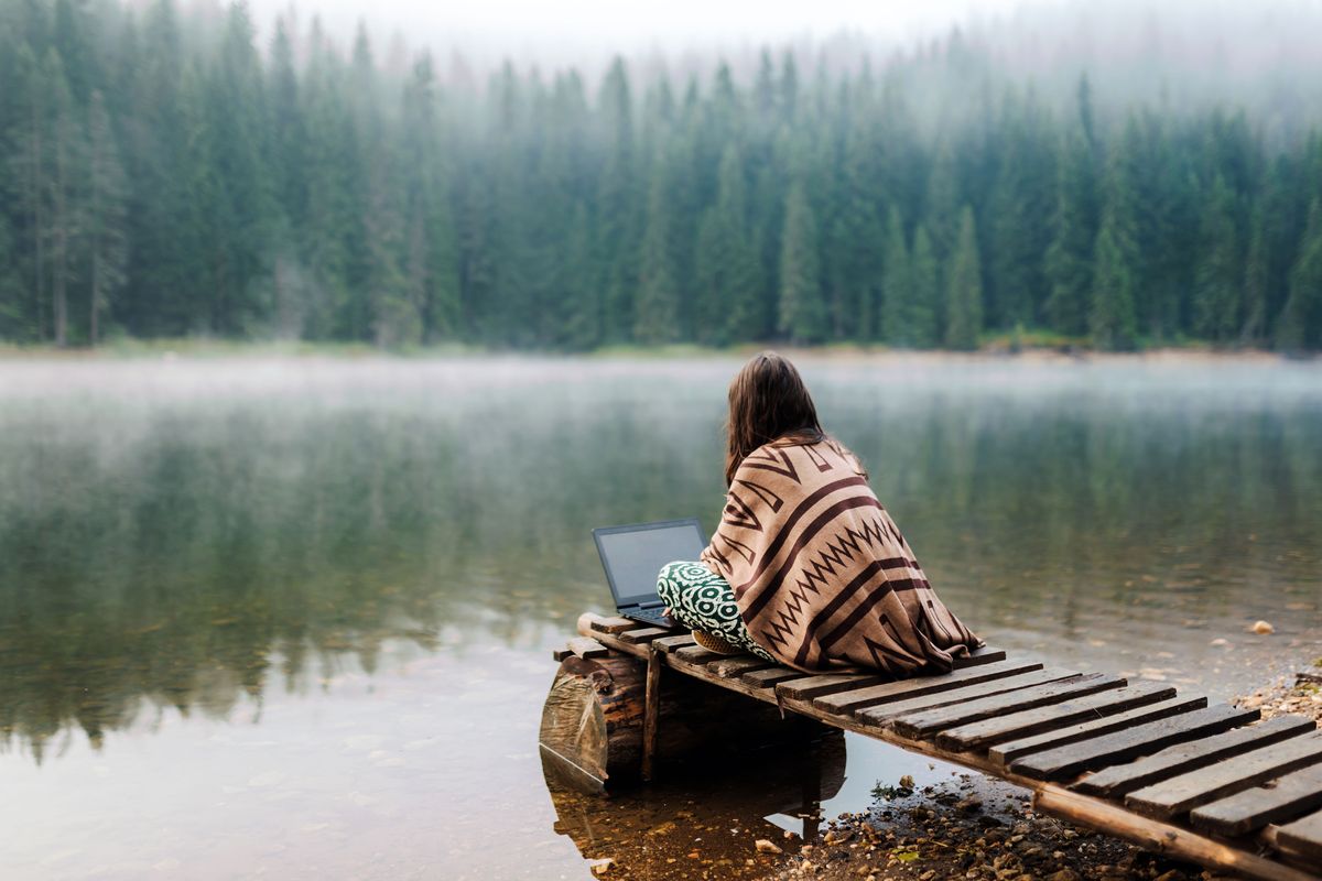 A woman works on her laptop while sitting on the dock of a lake.