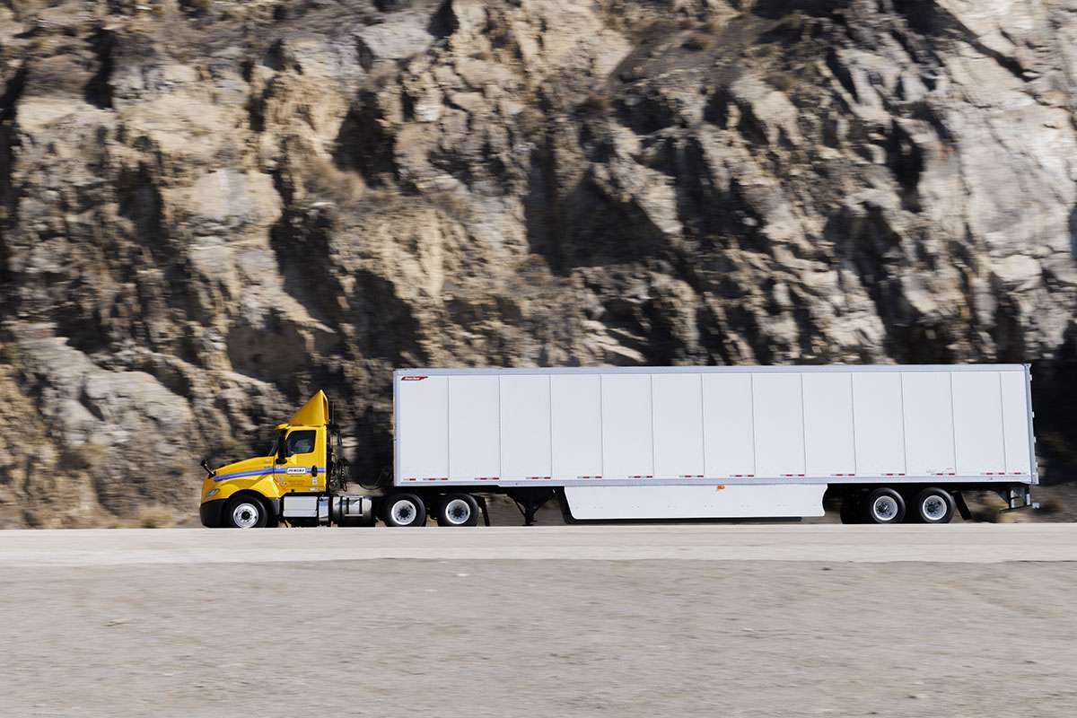 A yellow and white Penske semi-truck and trailer driving in front of a mountain of rocks.
