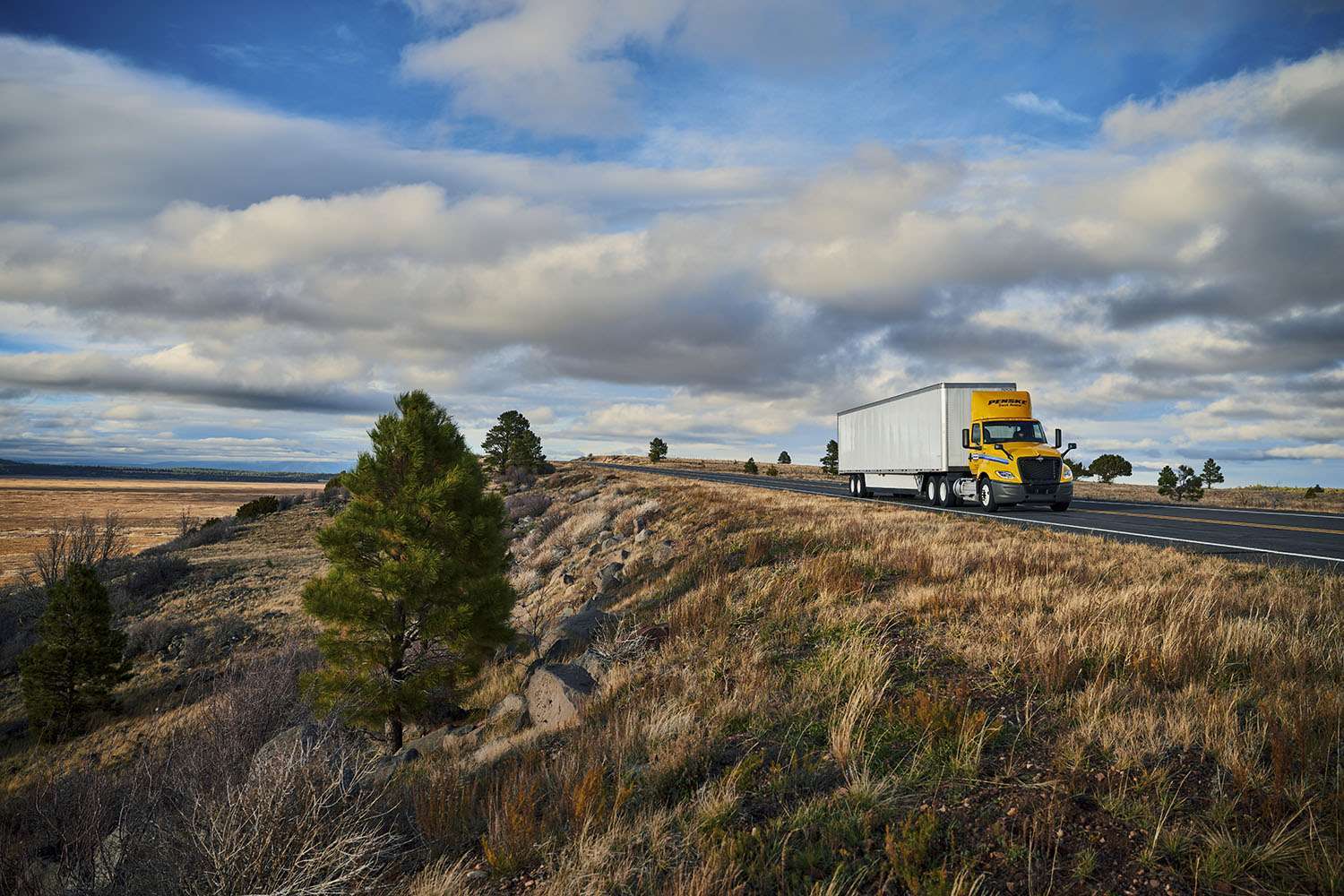 A yellow and white Penske semi-truck drives along a country road under a blue cloudy sky.