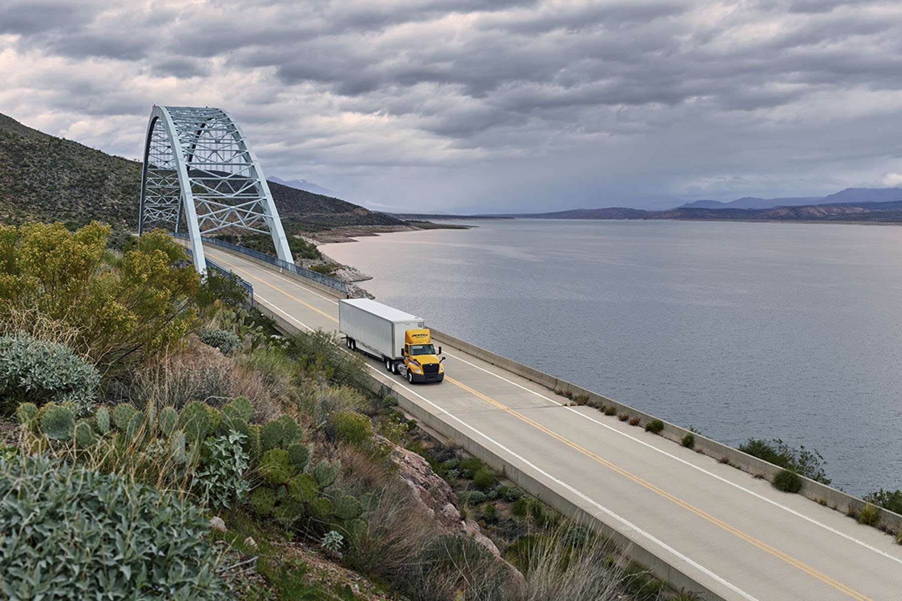 A yellow and white Penske semi-truck drives on a bridge over a body of water on a cloudy day.