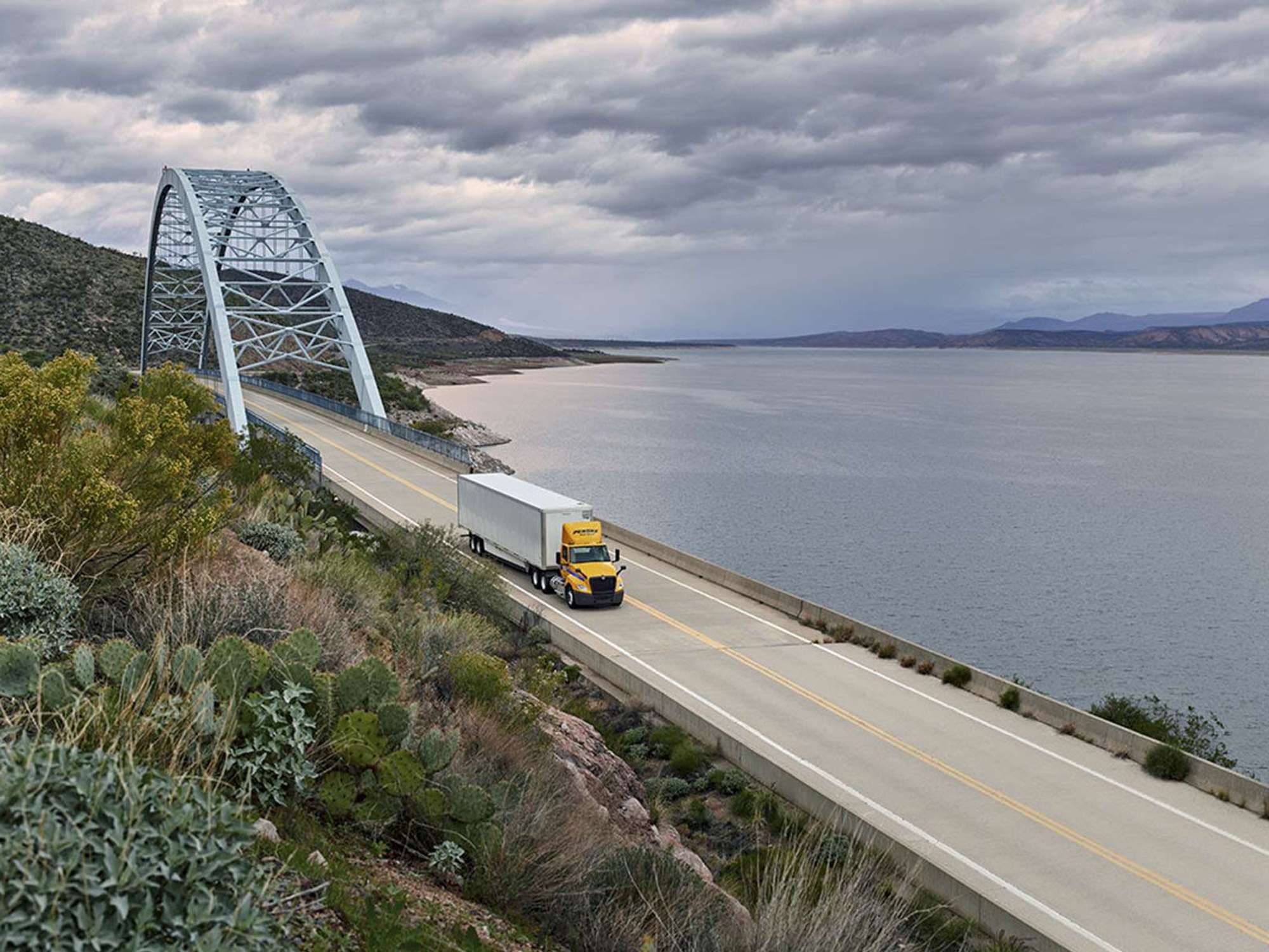 A yellow and white Penske semi-truck drives on a bridge over a body of water on a cloudy day.