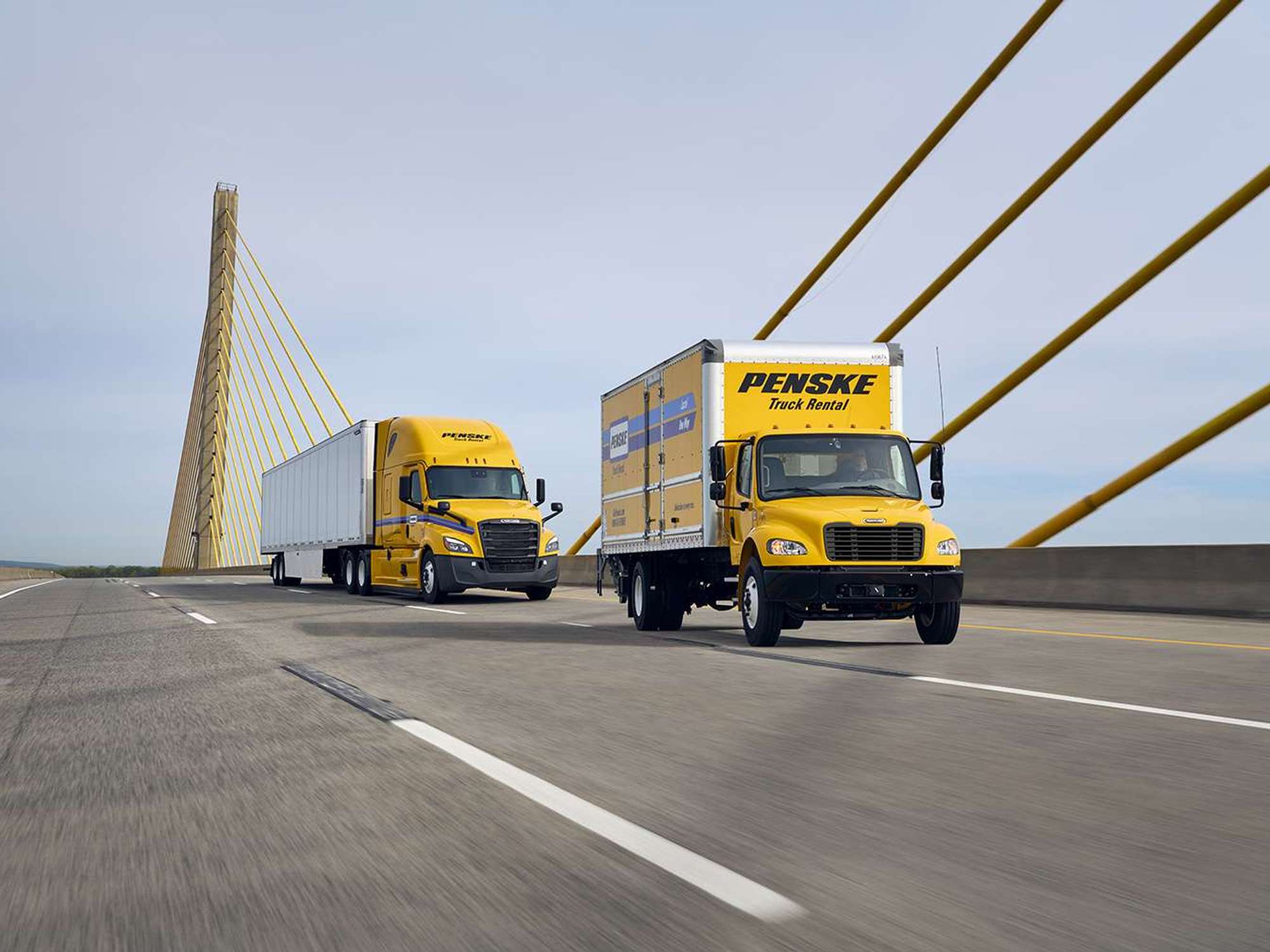 A yellow Penske box truck and yellow Penske semi-truck drive across a bridge.