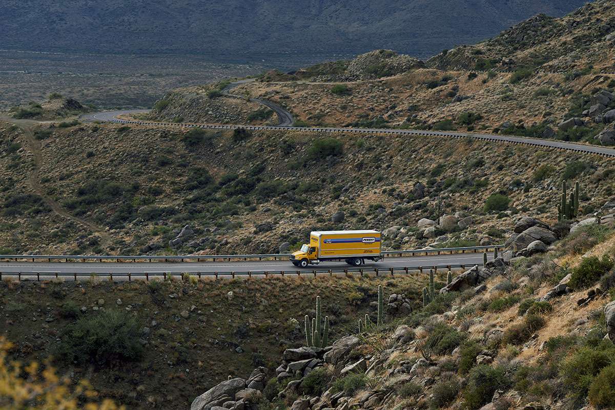 A yellow Penske box truck drives across a bridge in the rocky desert.