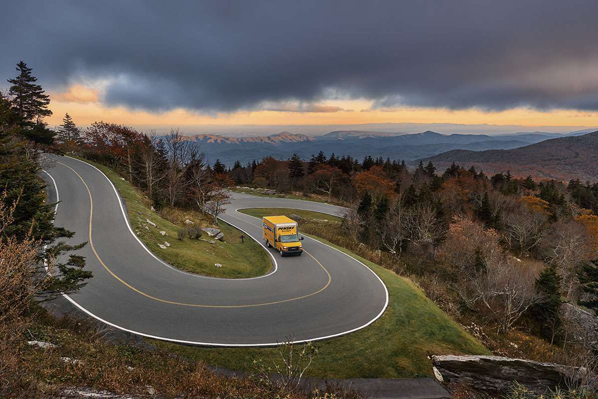 A yellow Penske box truck drives on a curvy road in the mountains under a dark cloudy sky.