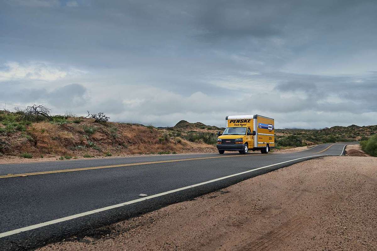 A yellow Penske box truck drives up a desert road against a cloudy sky.