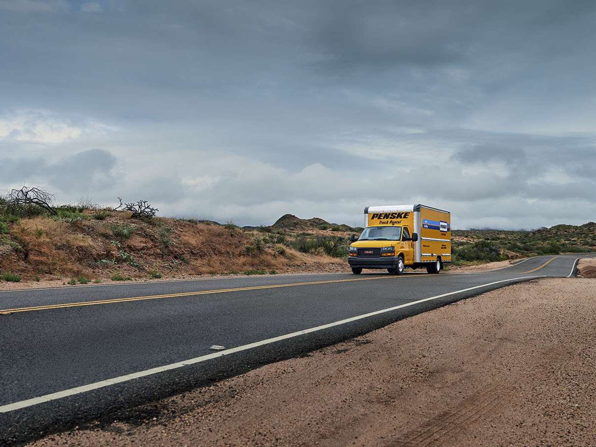A yellow Penske box truck drives up a desert road against a cloudy sky.