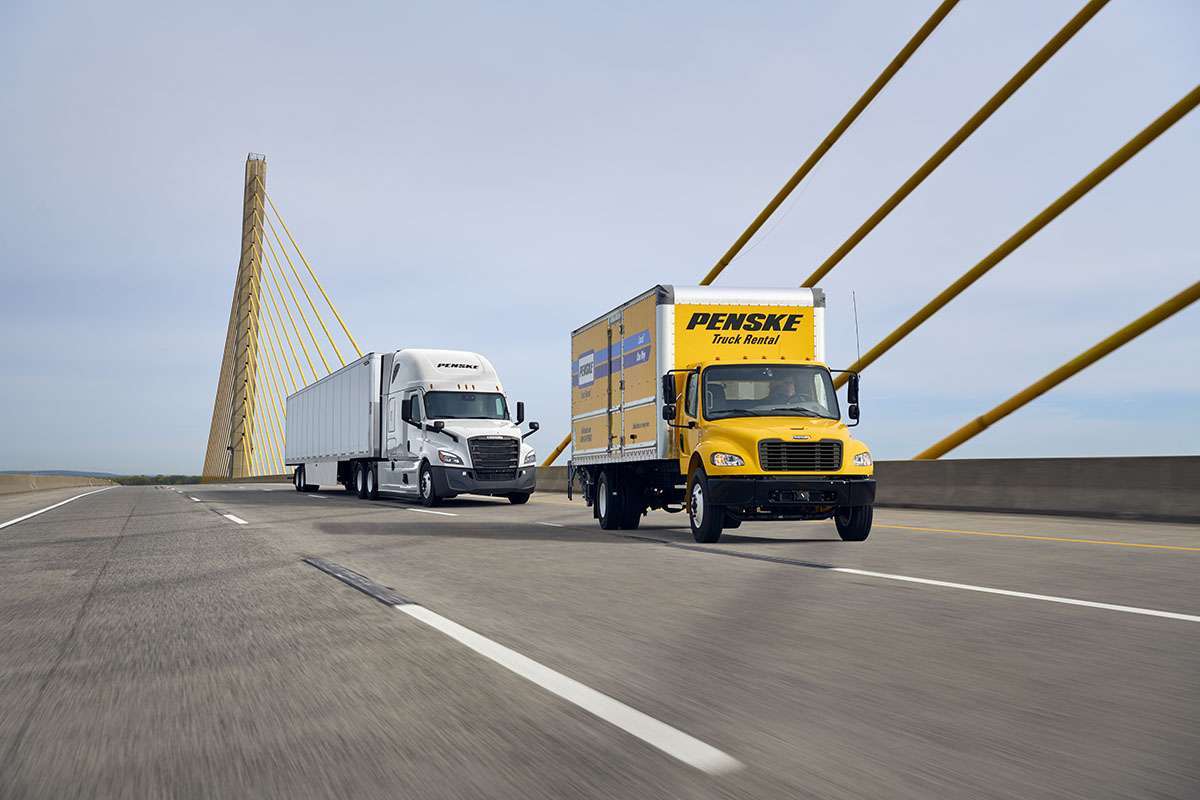 A yellow Penske branded box truck and a white semi-truck drive across a bridge.