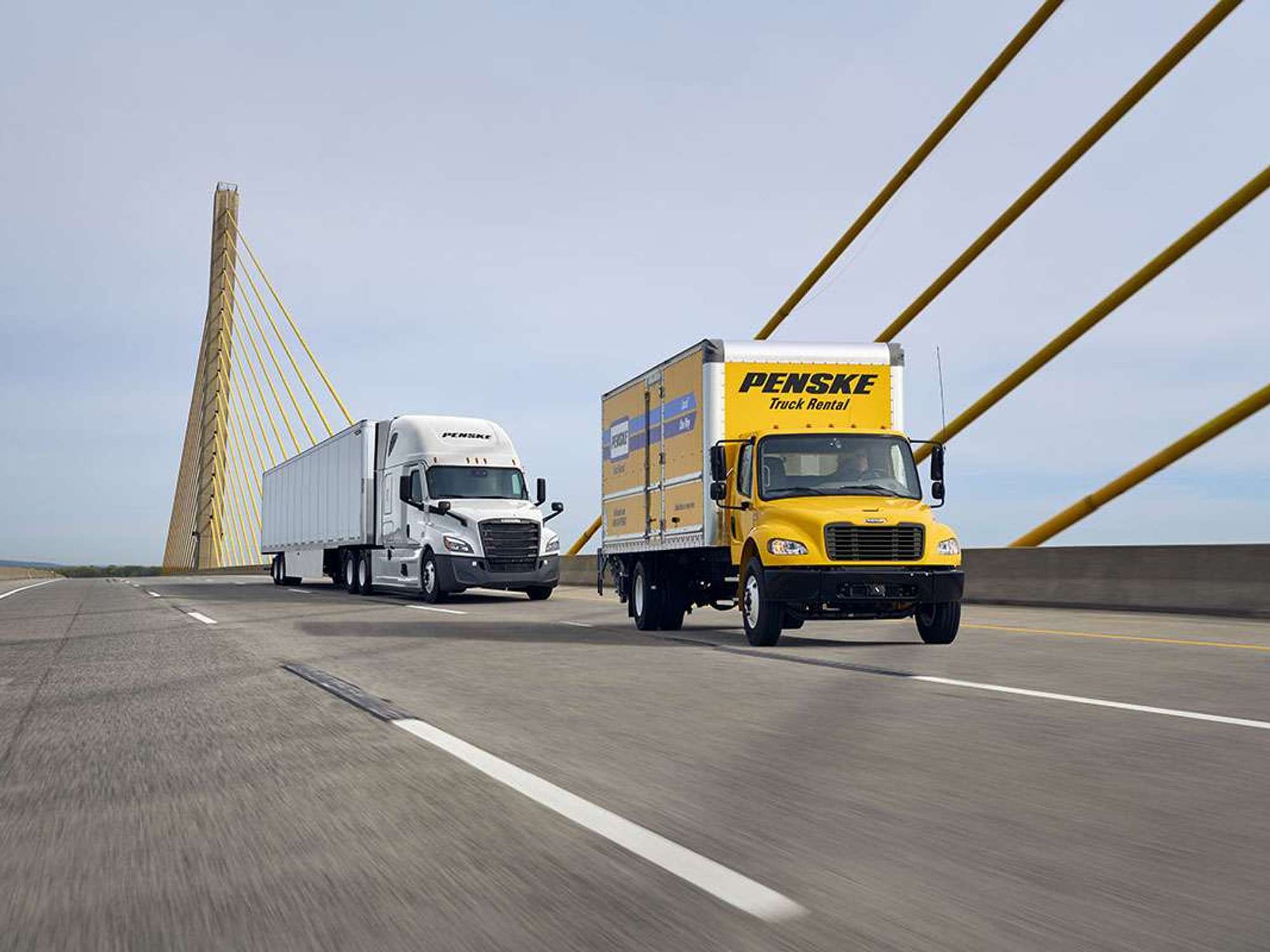 A yellow Penske branded box truck and a white semi-truck drive across a bridge.