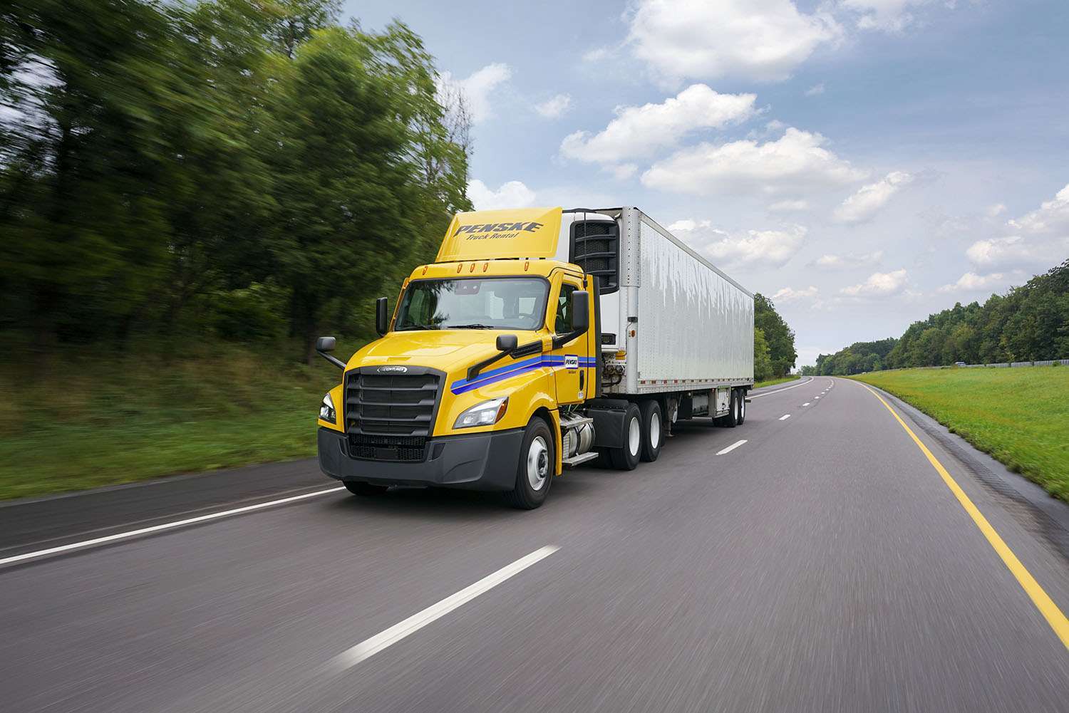 A yellow Penske semi-truck and white trailer drives up a two-lane road surrounded by green trees and blue sky.