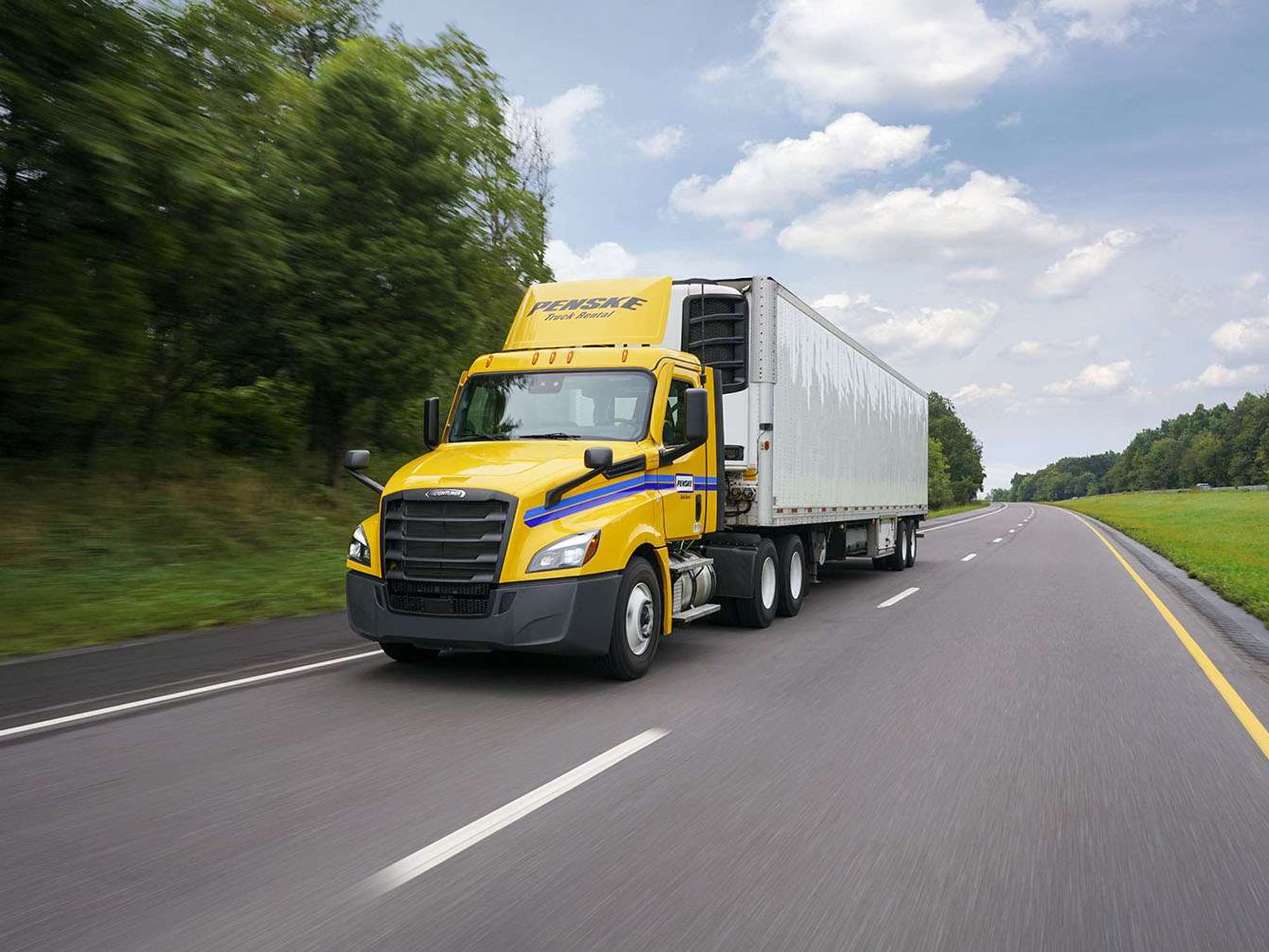 A yellow Penske semi-truck and white trailer drives up a two-lane road surrounded by green trees and blue sky.