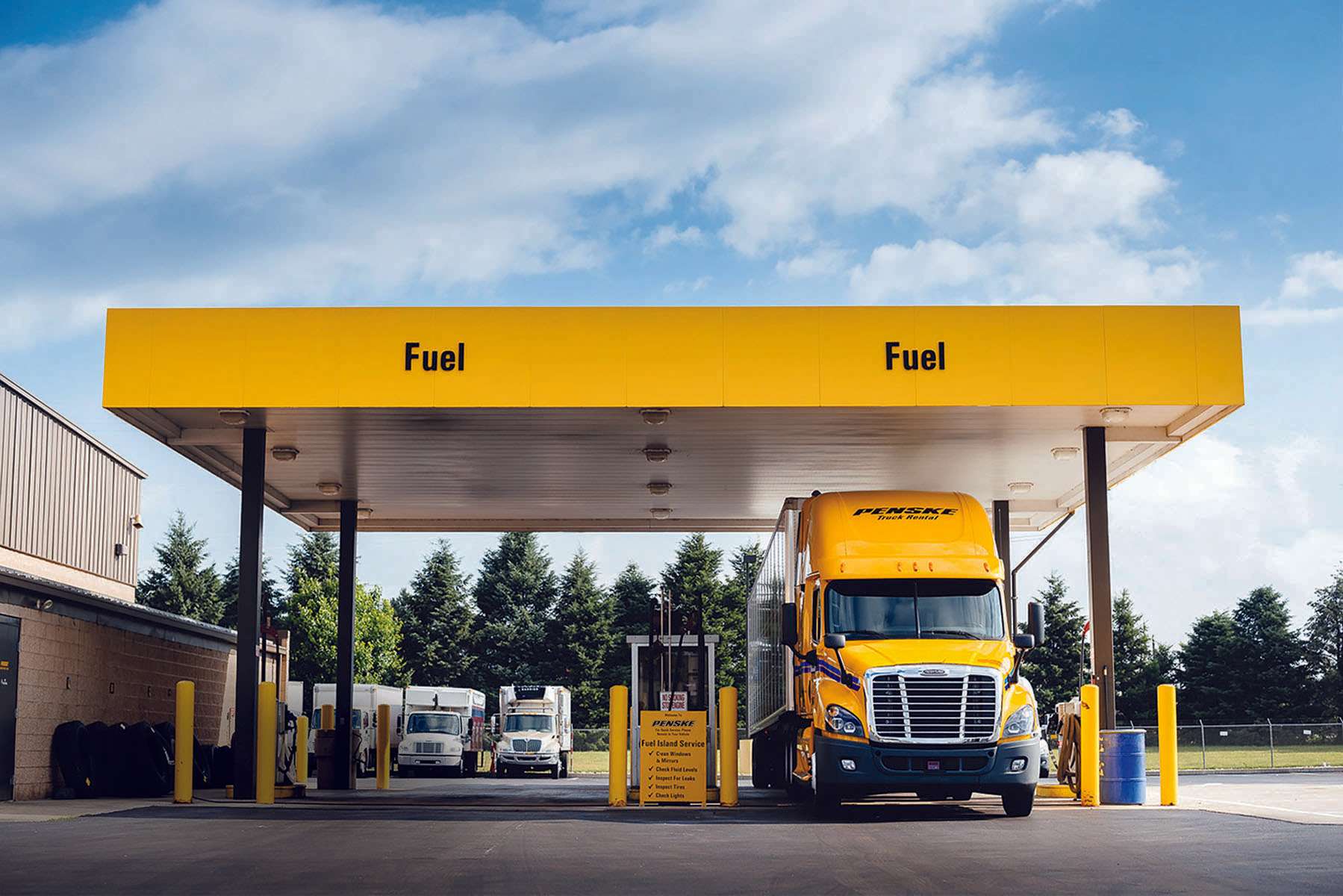 A yellow Penske semi truck is parked at a fuel pump underneath a yellow awning marked with the word "Fuel."