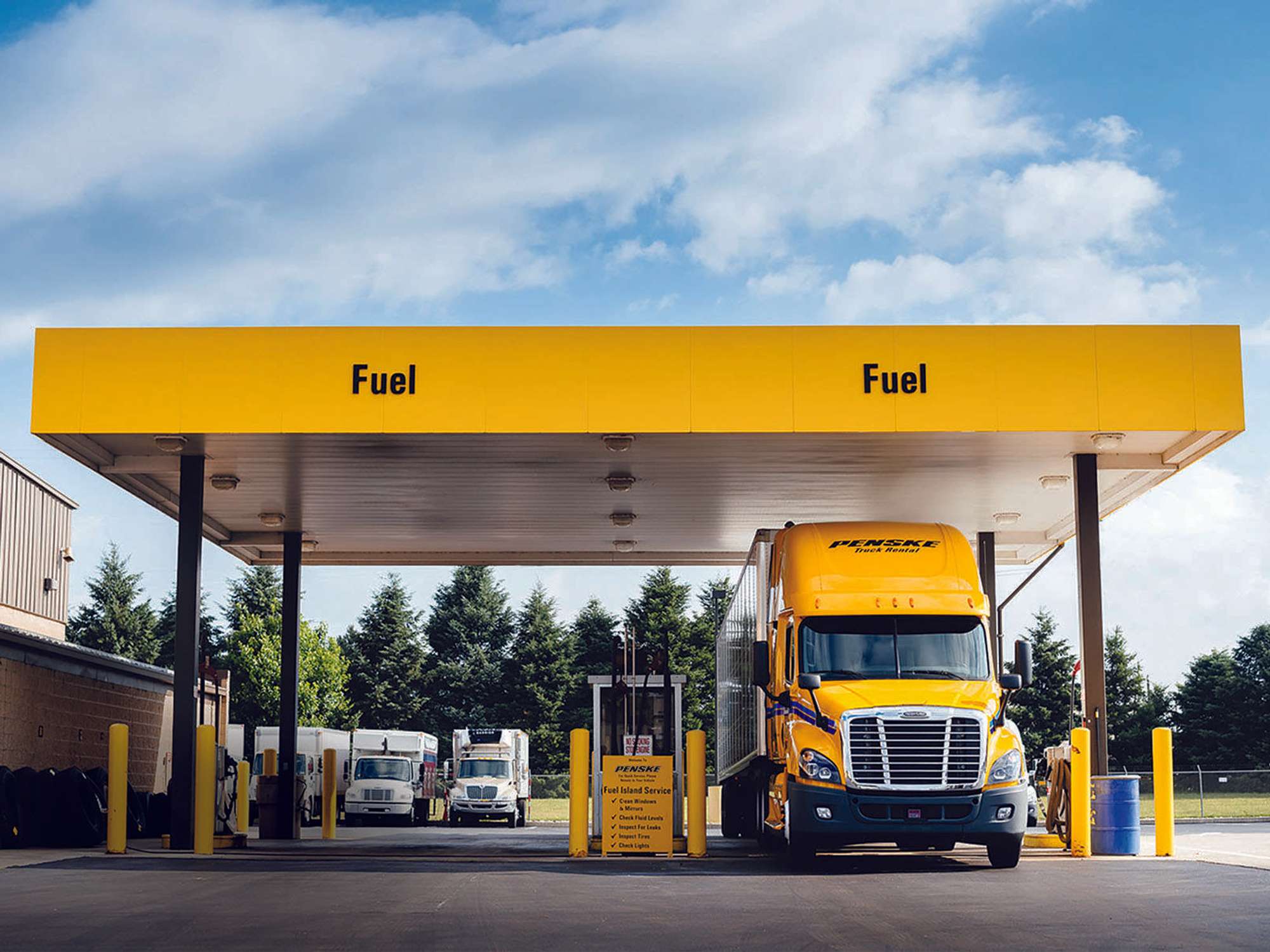 A yellow Penske semi truck is parked at a fuel pump underneath a yellow awning marked with the word "Fuel."
