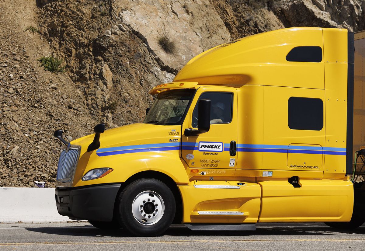 A yellow Penske semi-truck with white trailer attached sitting on a road near a mountain.