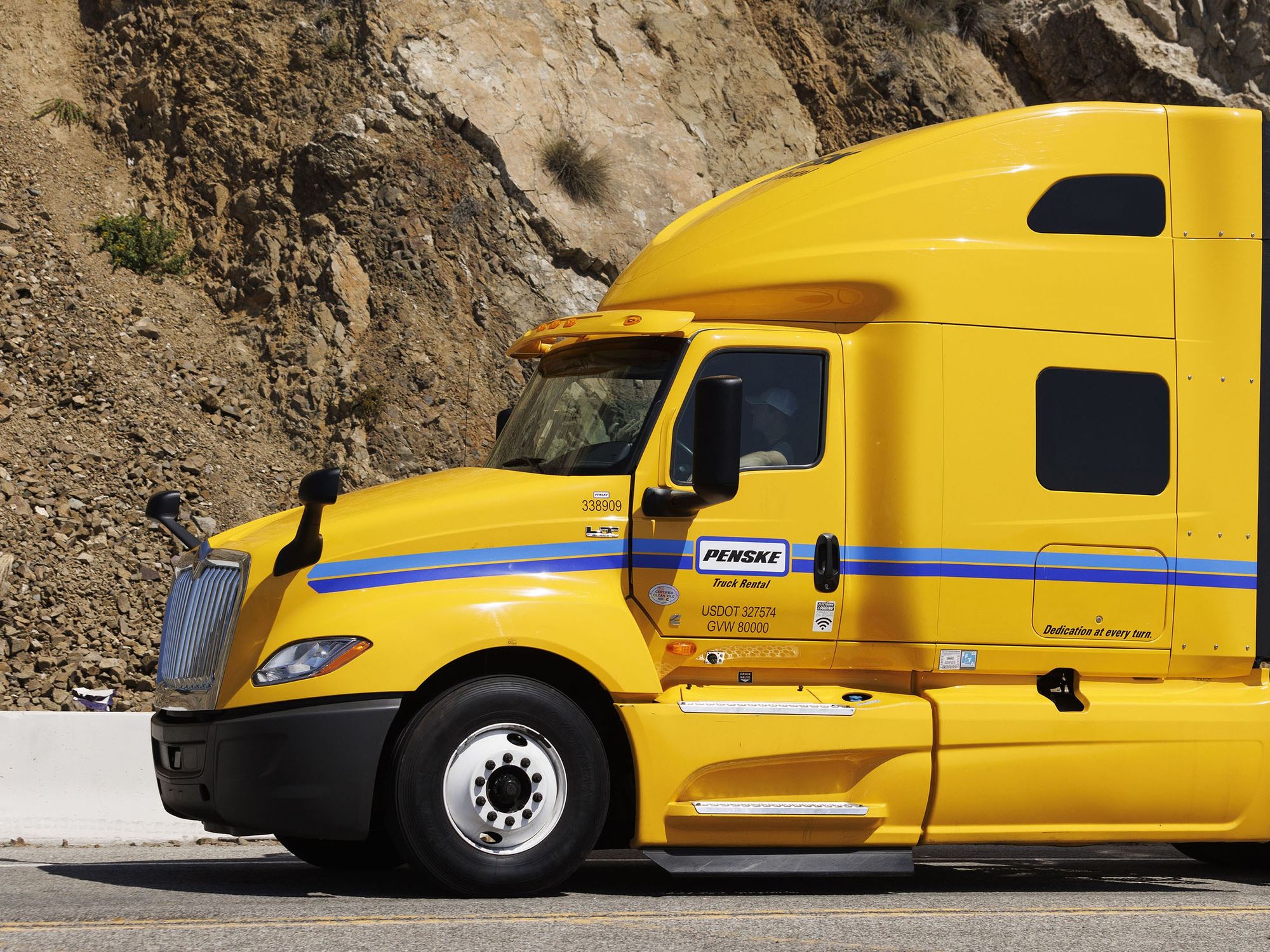 A yellow Penske semi-truck with white trailer attached sitting on a road near a mountain.