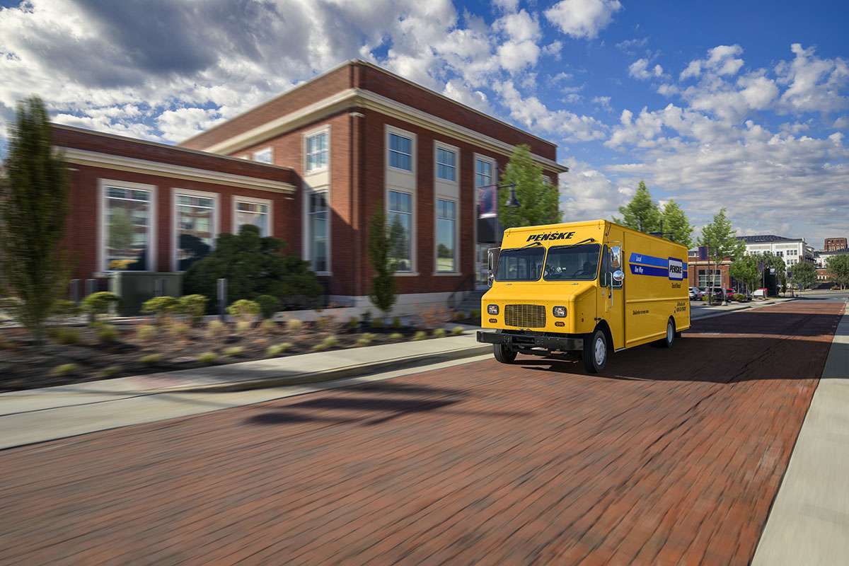 A yellow Penske step van drives up a brick street under a blue sky next to a brick building.