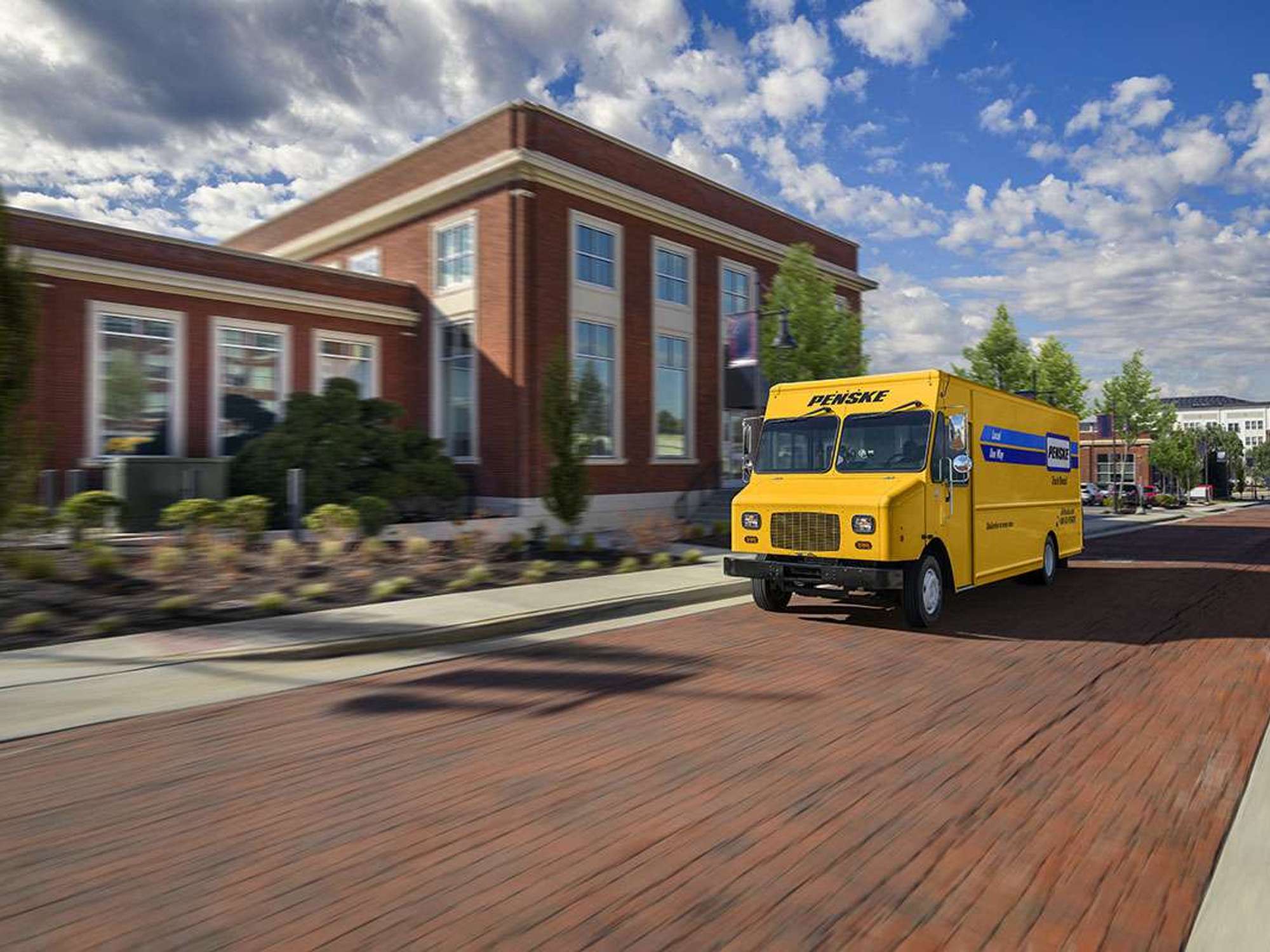 A yellow Penske step van drives up a brick street under a blue sky next to a brick building.