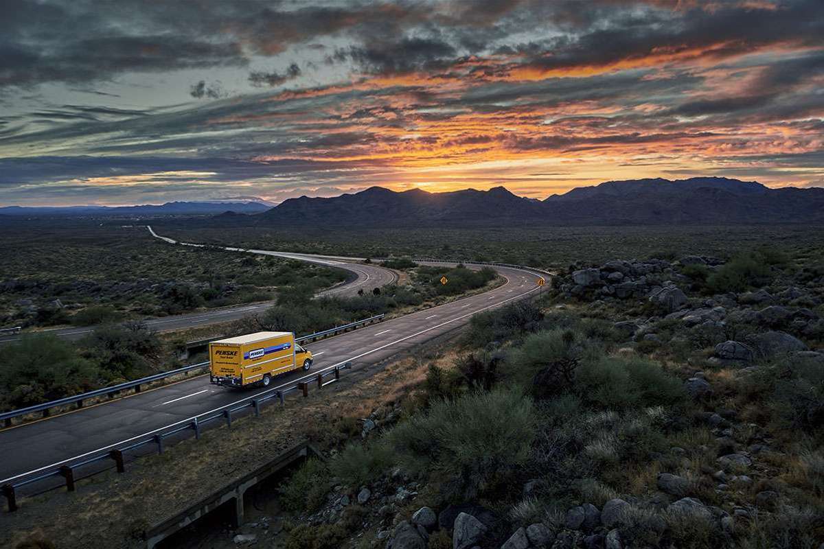 A yellow Penske truck drives up a road during sunset in the mountains.