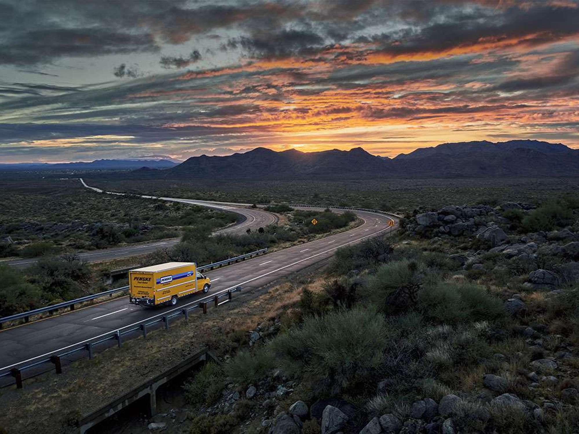 A yellow Penske truck drives up a road during sunset in the mountains.