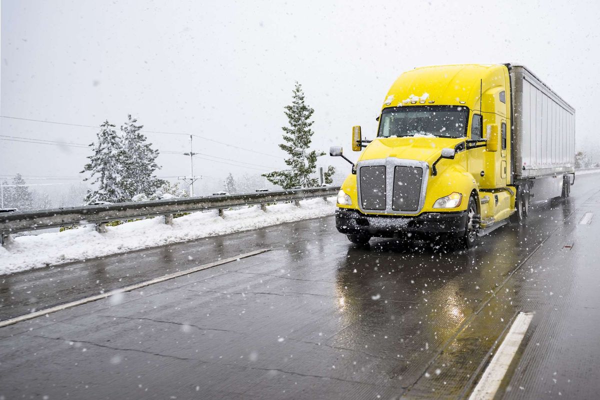 A yellow semi-truck with a silver trailer driving in the snow.