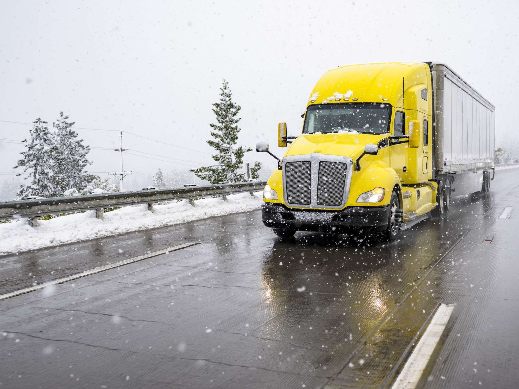 A yellow semi-truck with a silver trailer driving in the snow.