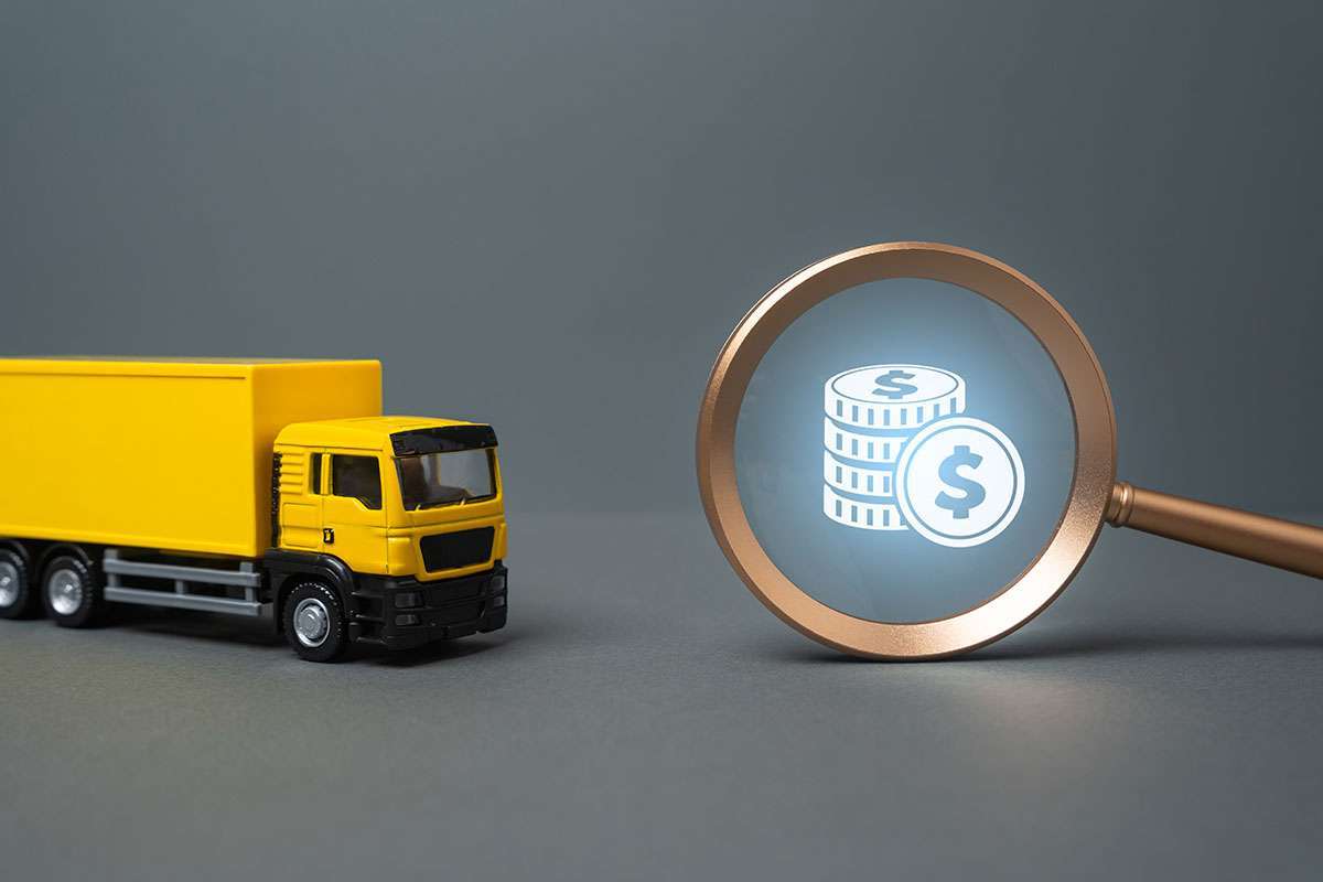 A yellow toy cabover truck sits next to a magnifying glass with a money icon inside.