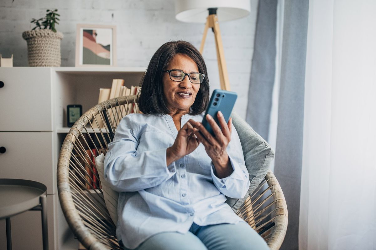 An older woman sits in a chair smiling while looking at her smartphone.