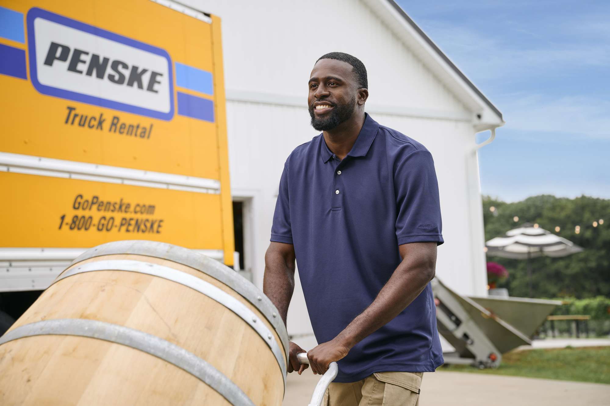 At a winery, a man pushes a wine barrel after unloading from a yellow Penske truck.