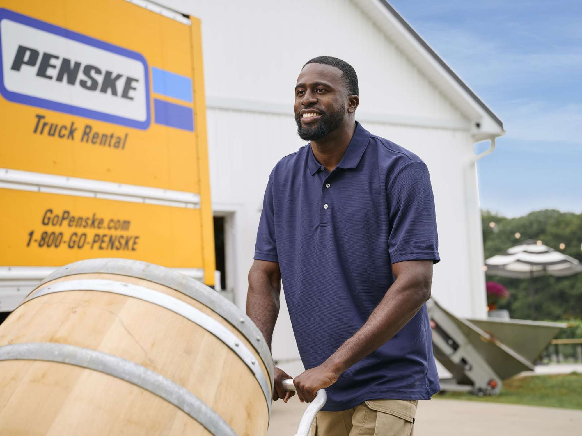 At a winery, a man pushes a wine barrel after unloading from a yellow Penske truck.