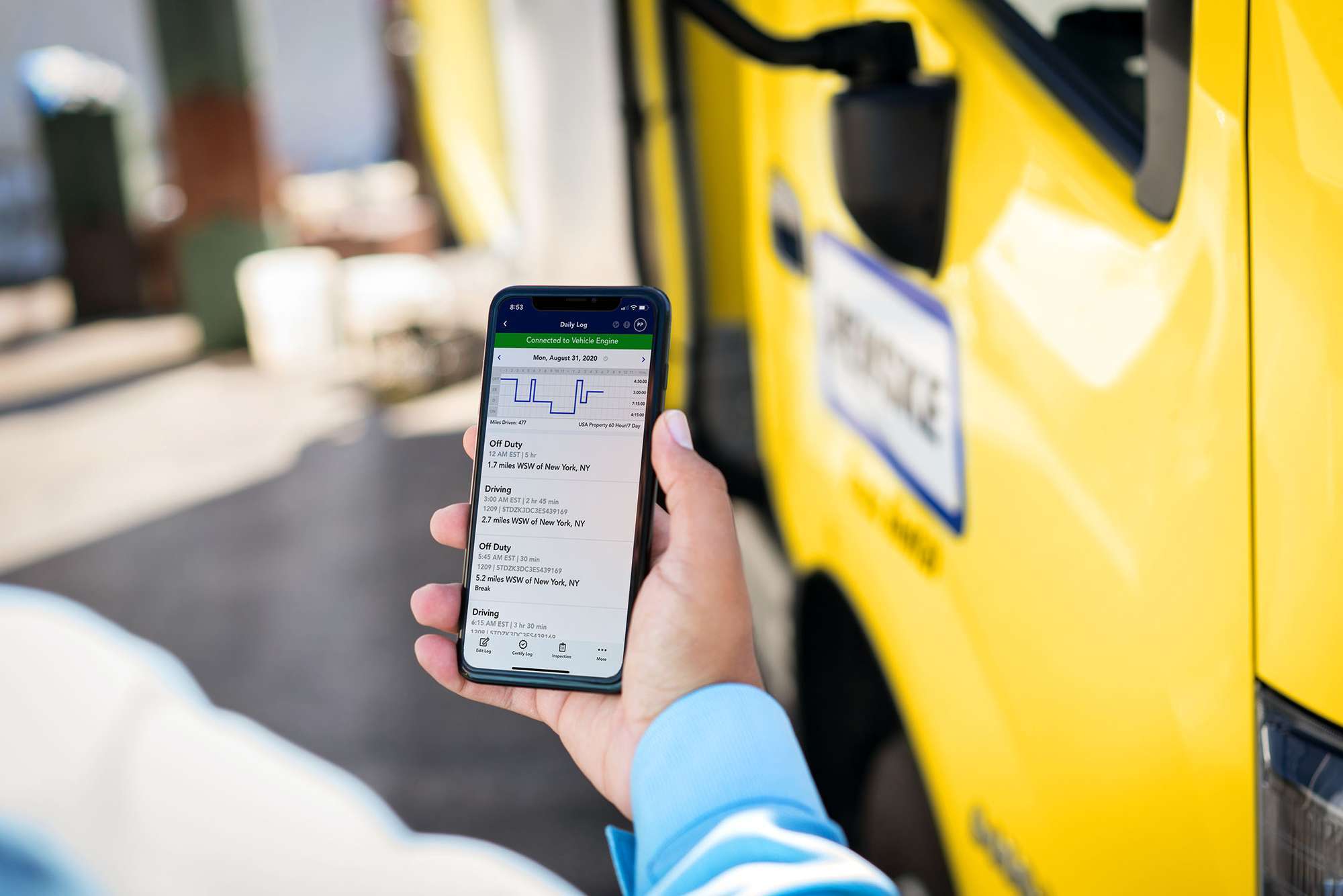 Closeup of a hand holding a cellphone with an hours of service log on the screen and a yellow Penske truck in the background.