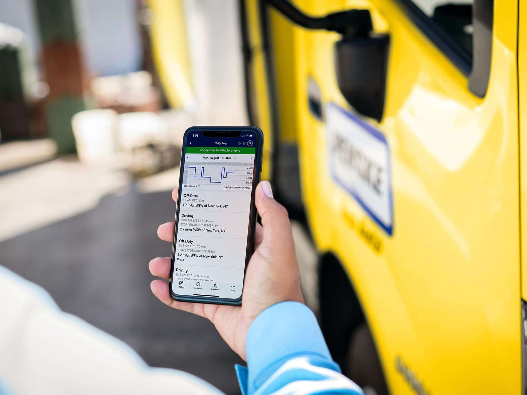 Closeup of a hand holding a cellphone with an hours of service log on the screen and a yellow Penske truck in the background.