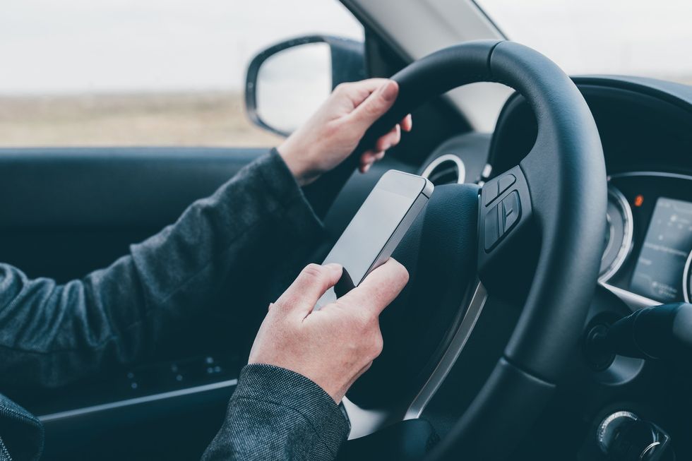 Closeup of a person's hands on the steering wheel of a car while also holding a smartphone. 