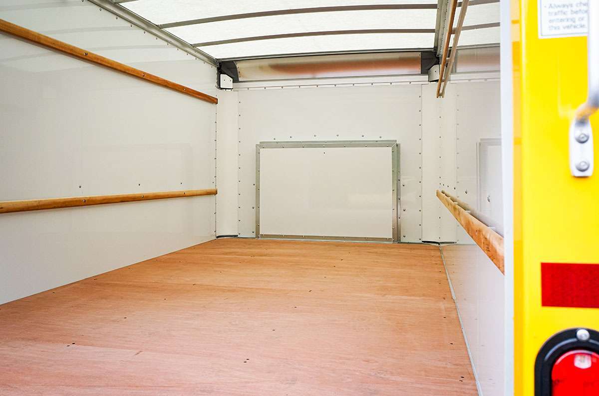 The inside back of an empty Penske box truck with brown wood floor, white walls, and wooden cargo tie rails.