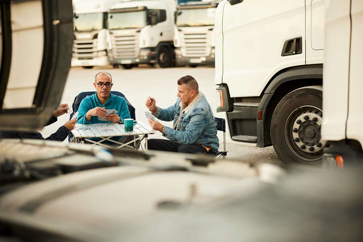 Three men sit around a table playing cards in a parking lot of semi-trucks.