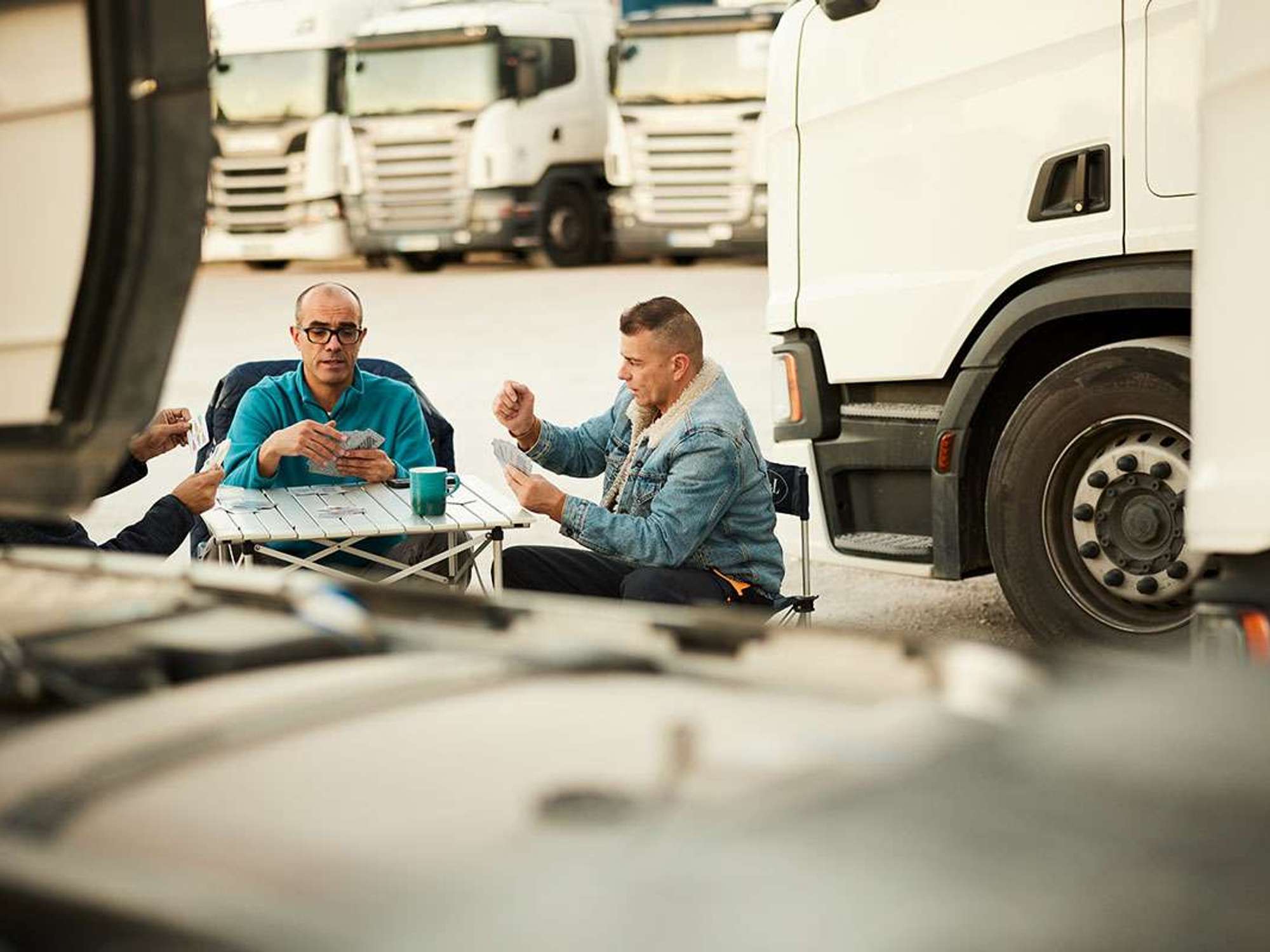 Three men sit around a table playing cards in a parking lot of semi-trucks.