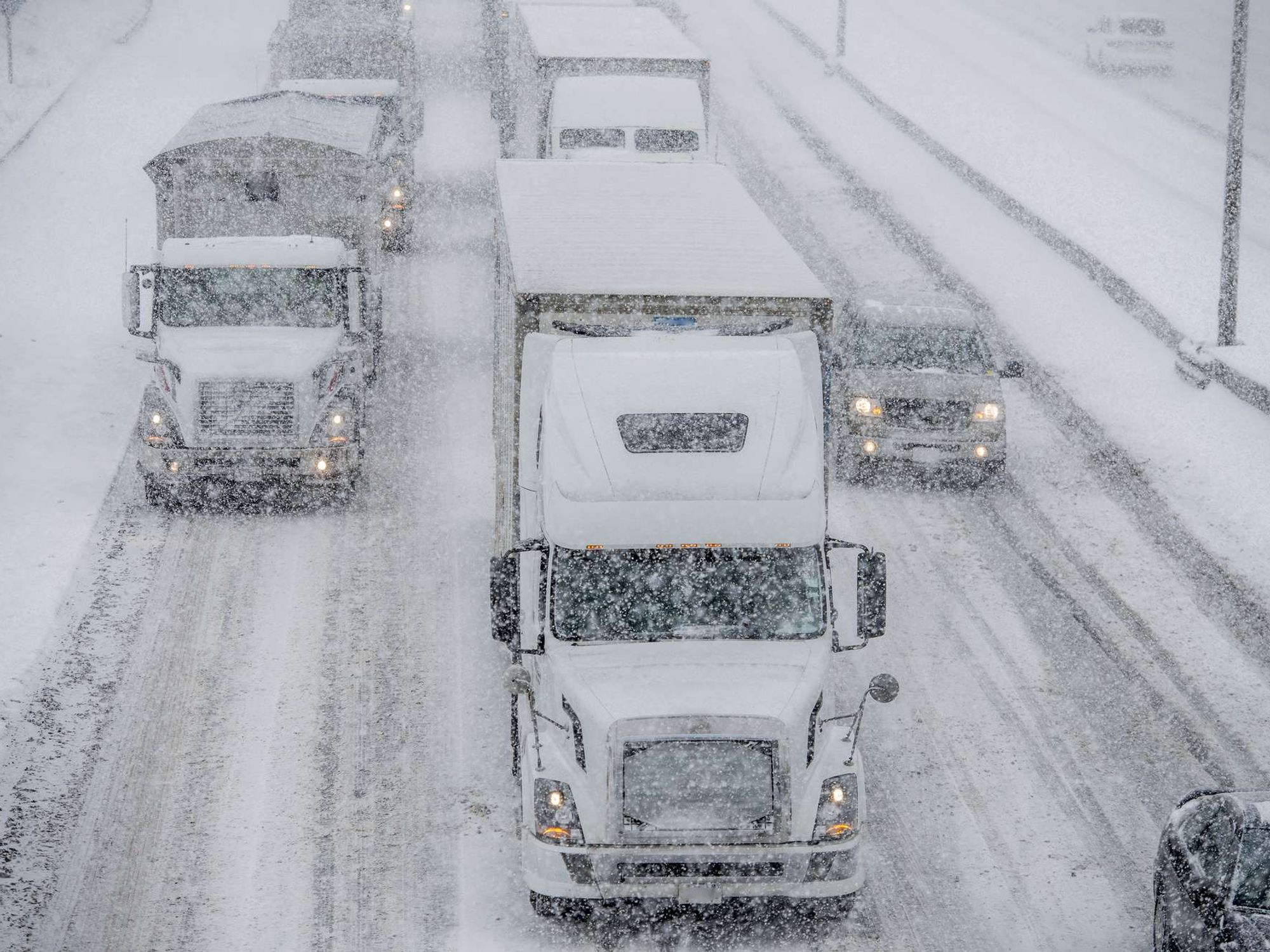 Trucks and cars drive on very snowy roads while it snows.