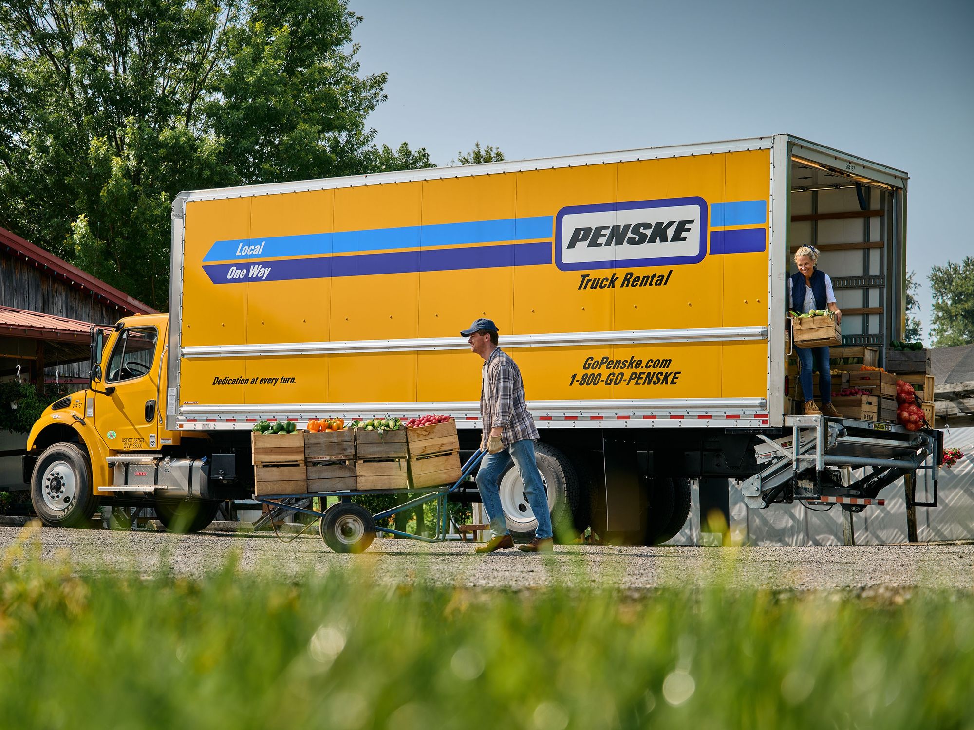 Two people deliver food in crates from the back of a yellow Penske truck to a farm store.