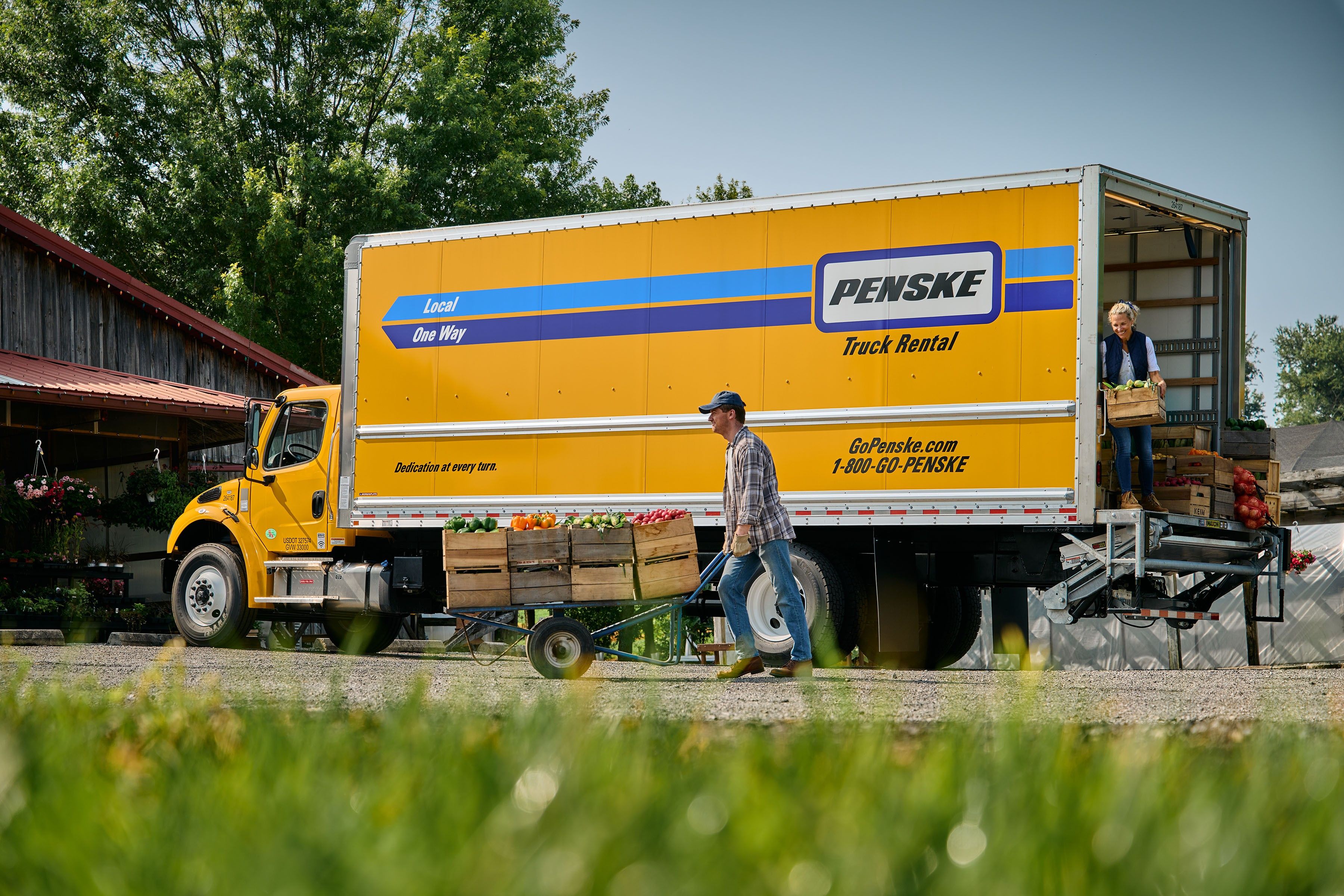 Two people deliver food in crates from the back of a yellow Penske truck to a farm store.