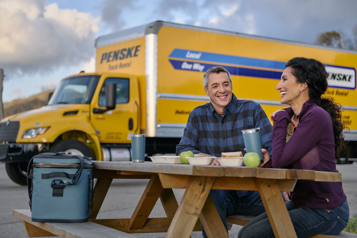 Two people enjoy a picnic lunch next to their yellow Penske moving truck.