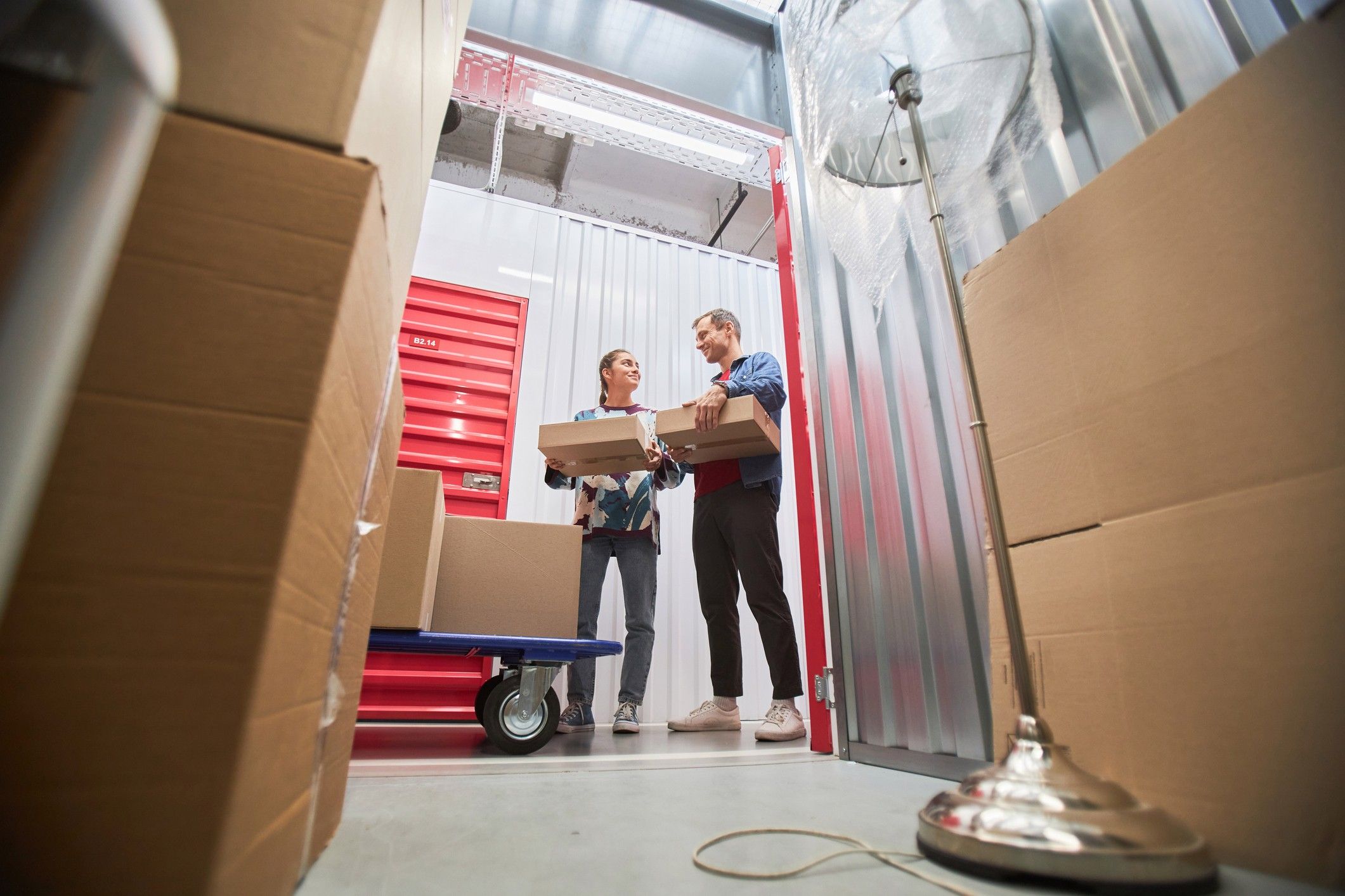 Two people holding boxes stand in front of an open storage unit surrounded by boxes and furniture.