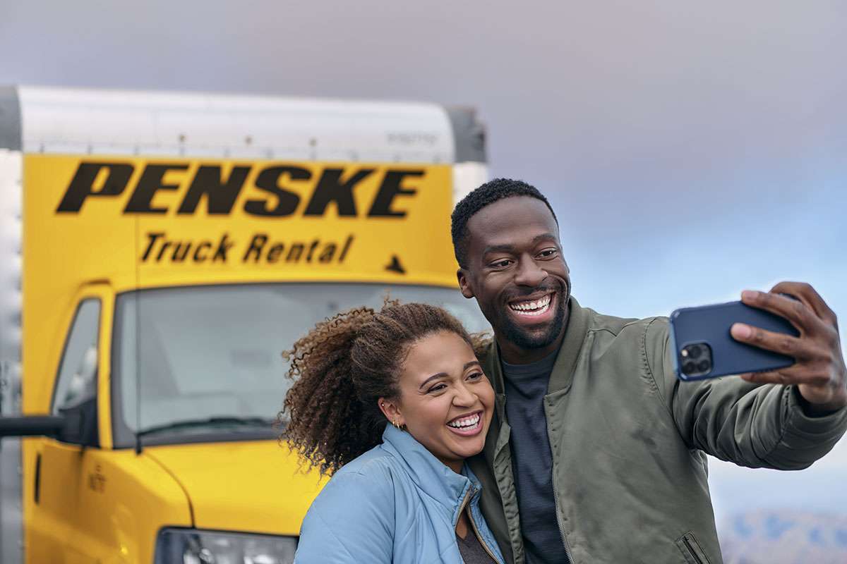 Two people take a selfie with a cell phone while standing in front of a yellow Penske truck.