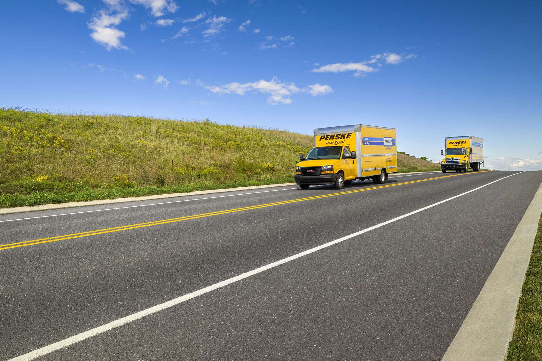 Two yellow Pensek rental trucks drive up a road on a sunny day.