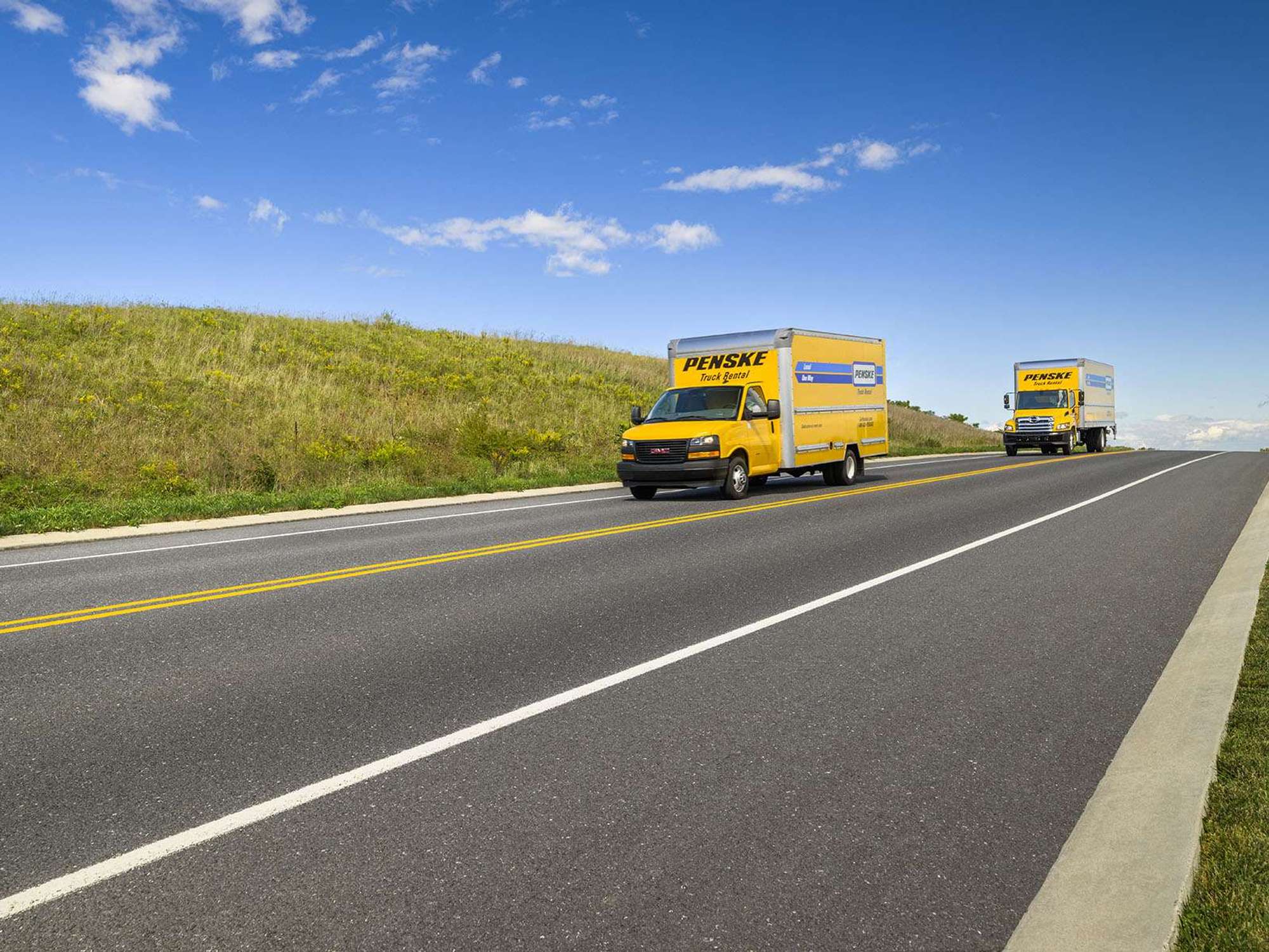 Two yellow Pensek rental trucks drive up a road on a sunny day.