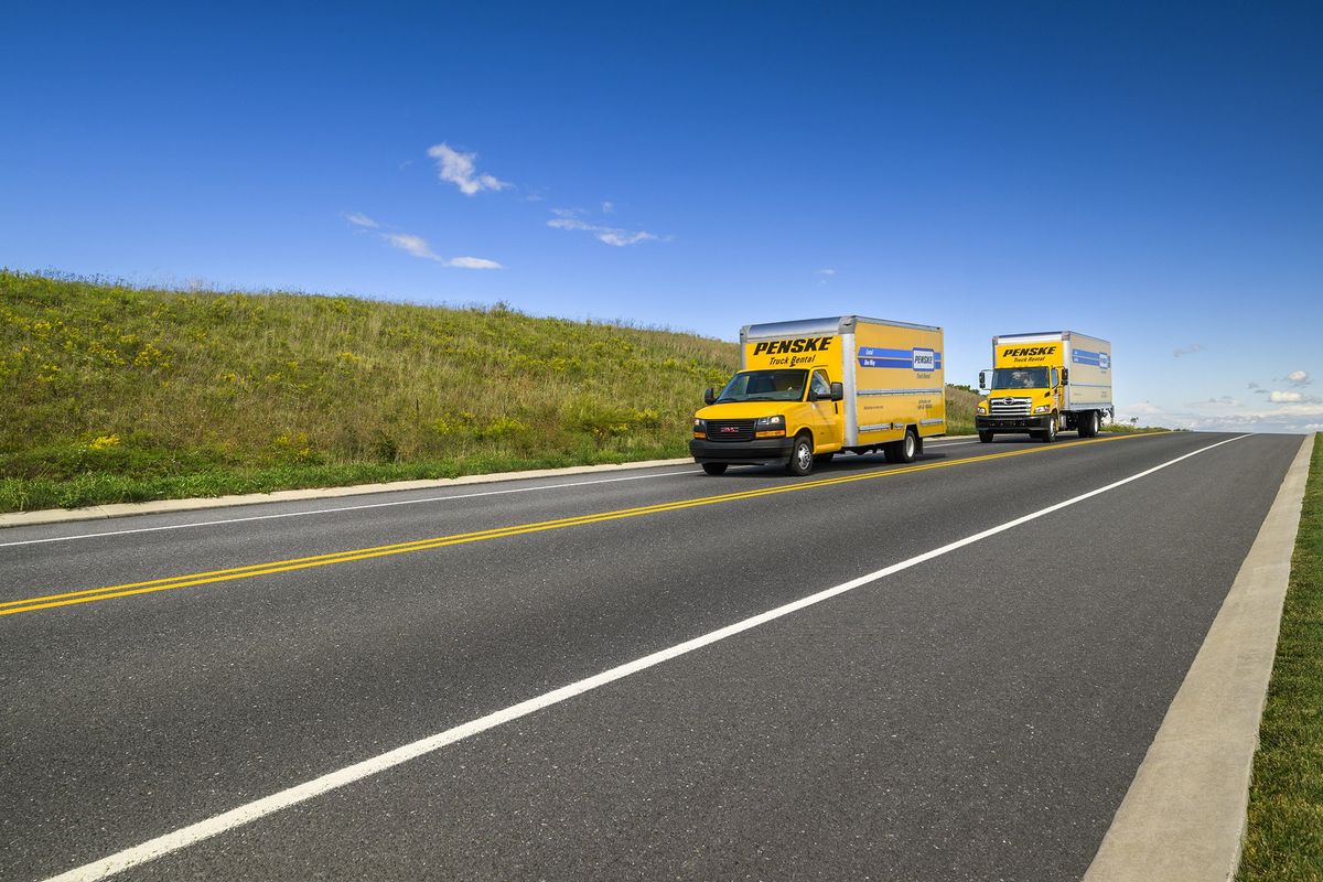 Two yellow Penske moving trucks drive up an empty road.