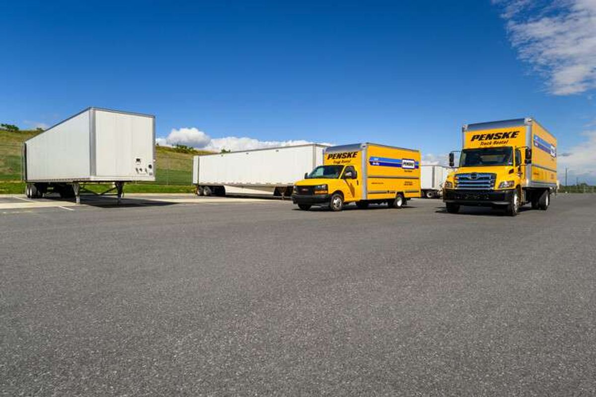 Two yellow Penske trucks parked in front of multiple white semi-trailers.