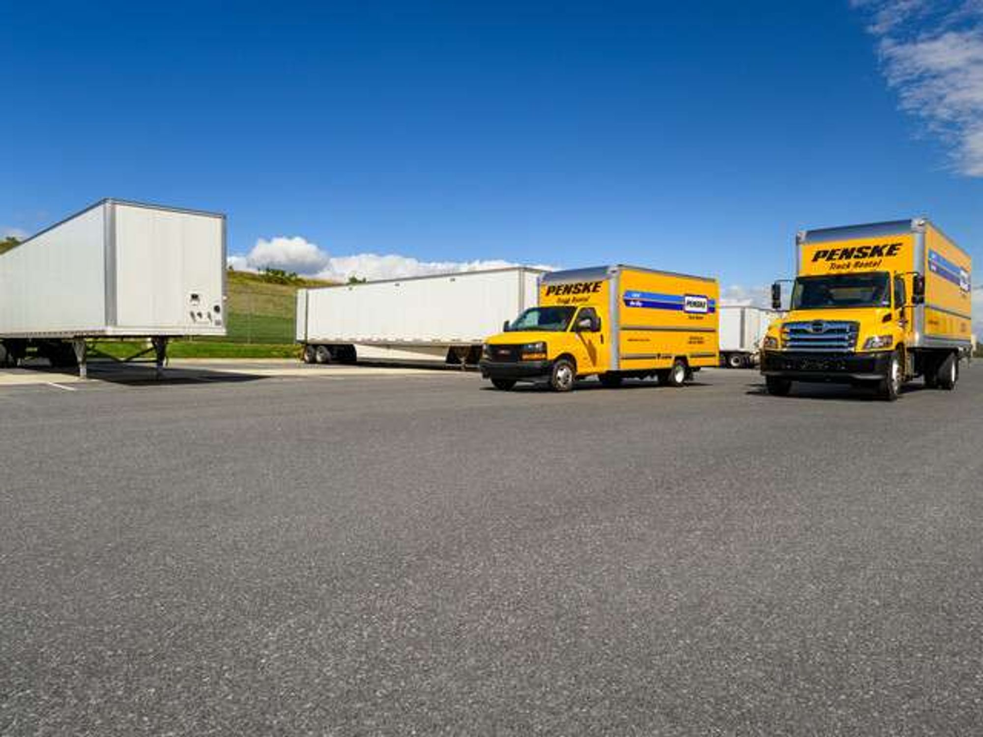 Two yellow Penske trucks parked in front of multiple white semi-trailers.