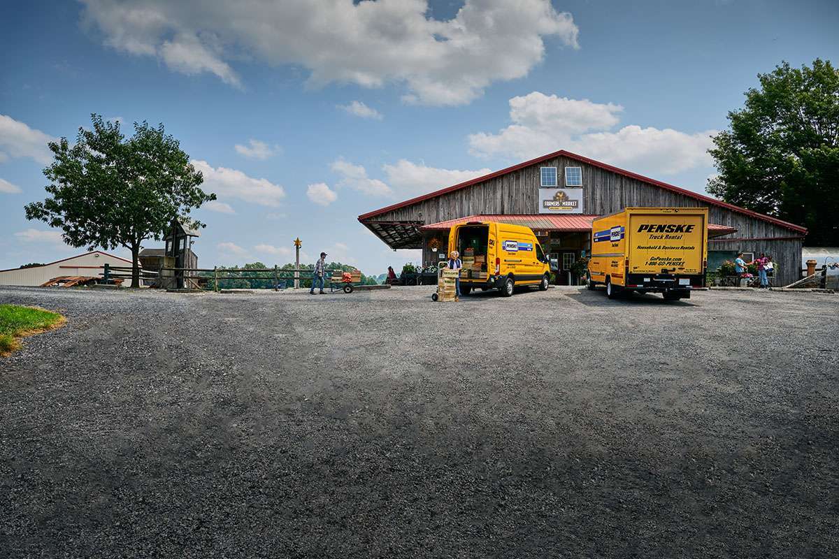 Two yellow Penske trucks parked outside a farmer's market unloading cargo.