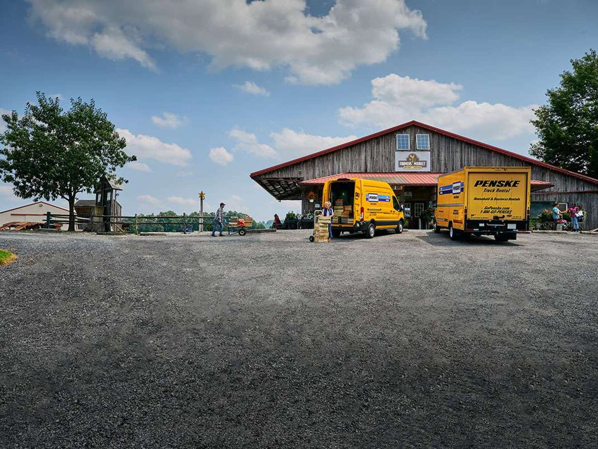 Two yellow Penske trucks parked outside a farmer's market unloading cargo.